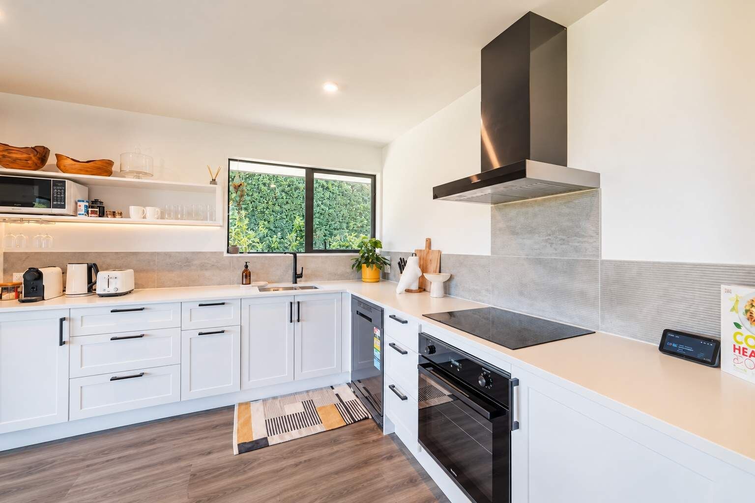 Une cuisine moderne avec des meubles blancs, un four encastré et une fenêtre donnant sur un espace verdoyant.