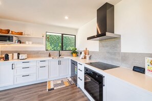 A modern kitchen with white cabinetry, a built-in oven, and a window overlooking greenery. - Hamptons Cottage | Hot Tub + Fireplace (Prebbleton)
