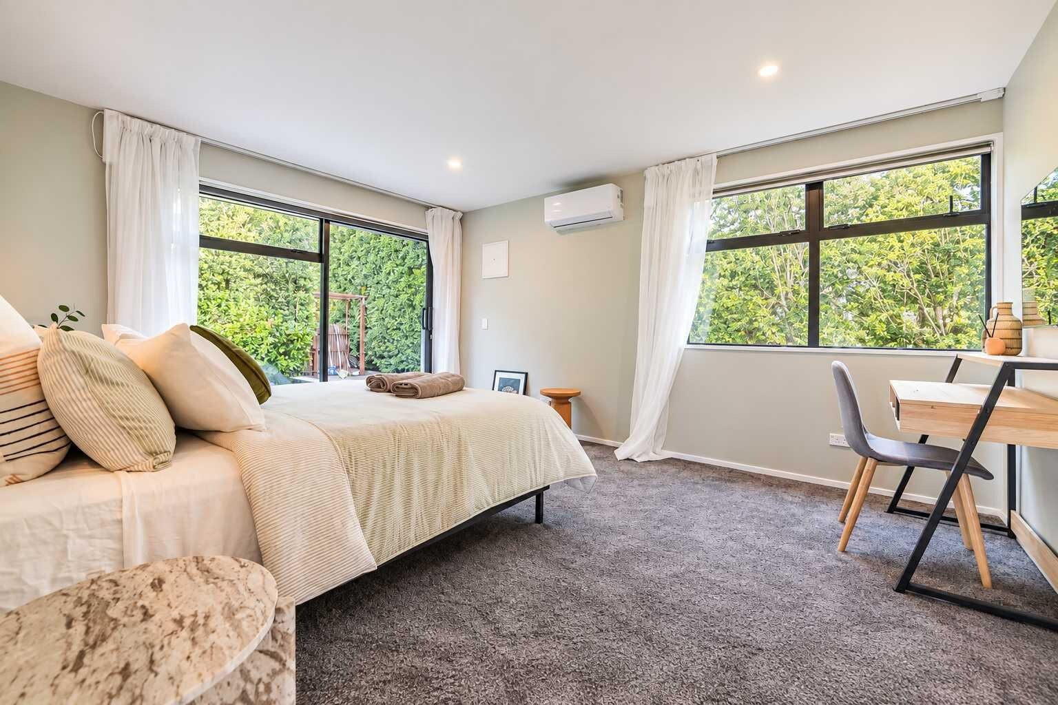 Une chambre avec un lit, un bureau et une chaise. Il y a des fenêtres avec des rideaux, une petite table et un climatiseur fixé au mur.