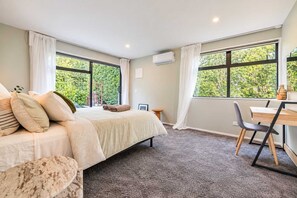 A bedroom with a bed, a desk, and a chair. There are windows with curtains, a small table, and a wall-mounted air conditioner. - Hamptons Cottage | Hot Tub + Fireplace (Prebbleton)
