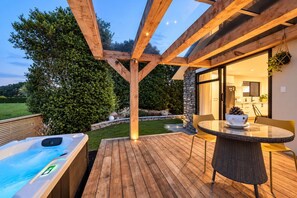 Une terrasse extérieure avec un jacuzzi, une table et des chaises, et une vue sur une maison dotée d’un mur en pierre et d’une porte en verre.