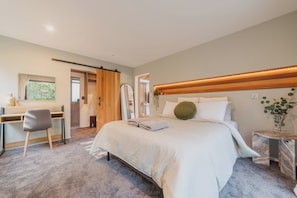 Un dormitorio con cama, escritorio y silla. Se ve vegetación por la ventana.