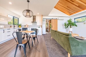 A modern kitchen with white cabinets, a dining area with a round table and chairs, a green sofa, and a wooden ceiling. - Hamptons Cottage | Hot Tub + Fireplace (Prebbleton)