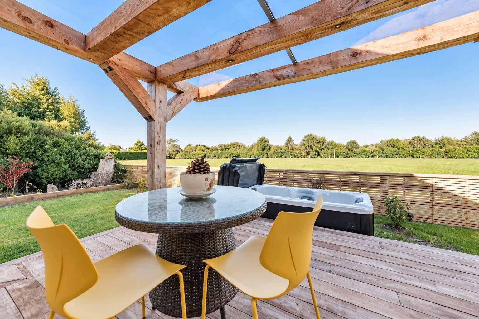 Une terrasse en bois avec une table ronde et deux chaises jaunes, un jacuzzi et une vue sur un champ verdoyant.