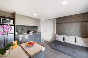 Une cuisine moderne équipée d’un four à micro-ondes, d’un évier et de placards, une table à manger avec un panier de fruits, et un canapé gris agrémenté de coussins à motifs.