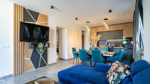 A modern living room with a blue sofa, a flat-screen TV mounted on a wooden panel, and a dining area with blue chairs and a table. - La Vista Azure (Sukosan)