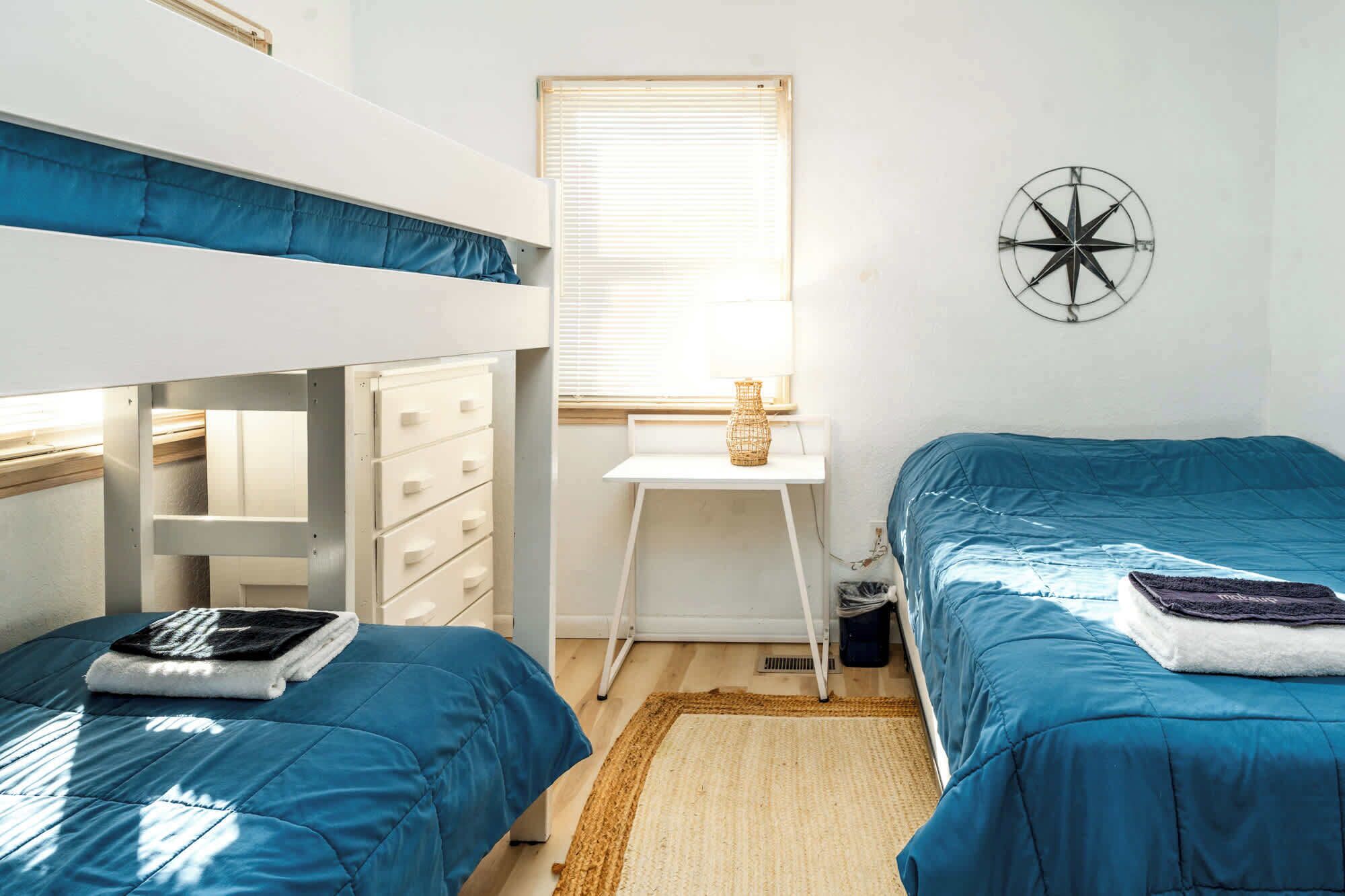 A dormitory room with bunk beds, a desk, a window with blinds, and a wall-mounted compass.