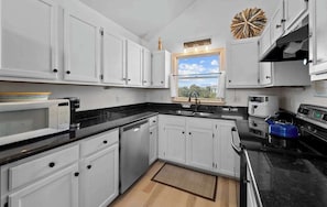 A modern kitchen with white cabinets, a black countertop, a window with a view, and a decorative wall piece. - Oceanside with Pool Hot Tub and Updated Interior! (Corolla)
