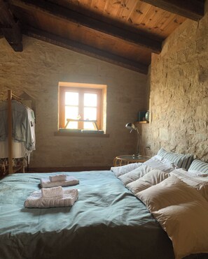 Une chambre rustique avec un grand lit, un plafond en bois, un mur en pierre et une fenêtre laissant entrer la lumière du soleil.