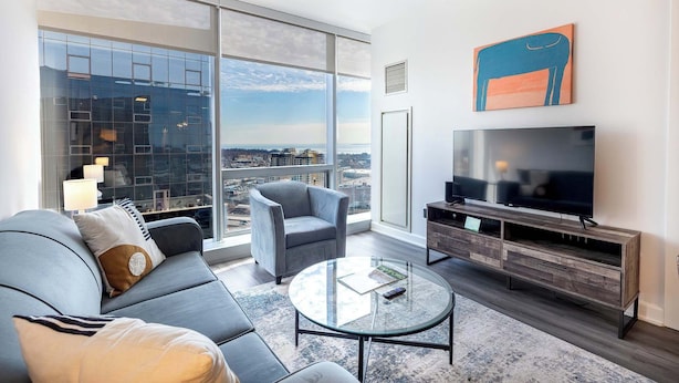 A modern living room with a large window, a sofa, a chair, a coffee table, and a TV. - Luxury Modern Apt with Pool (Stamford, CT)