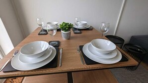 Une table à manger dressée avec de l’assiette blanche, des bols, des couverts et des verres à vin, agrémentée d’une petite plante en guise de centre de table.