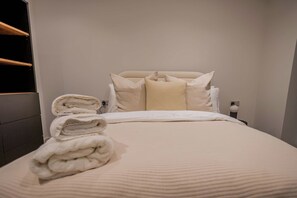 Una cama bien hecha con una toalla doblada a un lado, cabecero y mesita de noche con una lámpara.