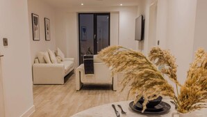 A modern living room with a sofa, a dining table with cutlery, and a vase of dried plants. - Bianco Suite 2BR with City Views (Greater Manchester)