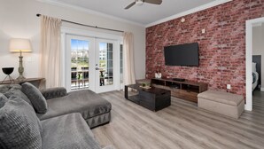 A modern living room with a brick wall, a flat-screen TV, a ceiling fan, and a large window with curtains. - First Floor Condo with Golf Course Views (Kissimmee)