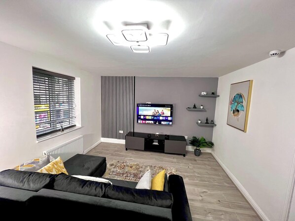 A modern living room with a flat-screen TV, a black sofa, a patterned rug, and a window with blinds.