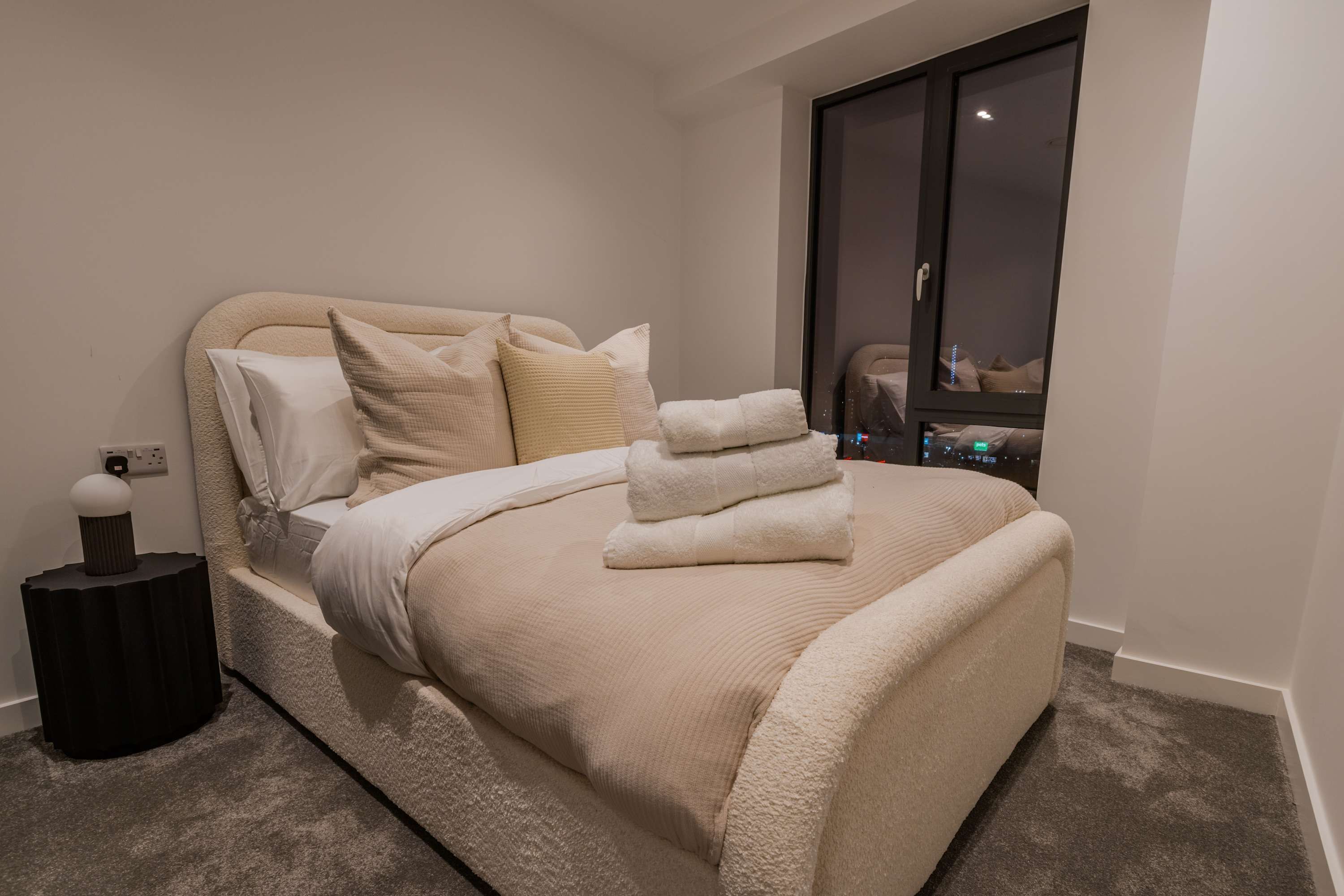 A neatly made bed with a beige comforter and pillows, a small black side table with a lamp, and a view of a balcony with a chair.