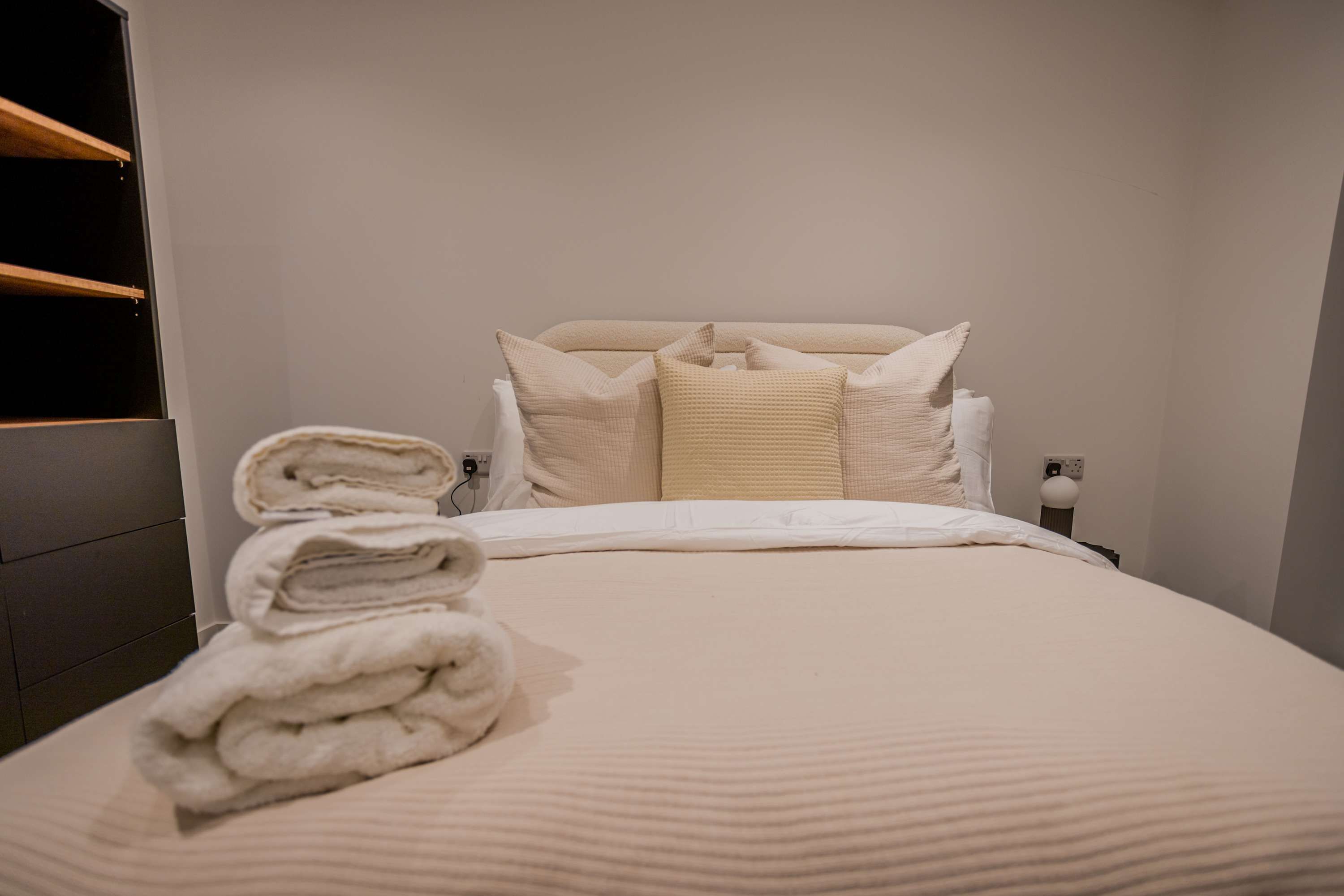 Un lit bien fait, avec une serviette pliée posée sur le côté, une tête de lit et une table de chevet sur laquelle se trouve une lampe.
