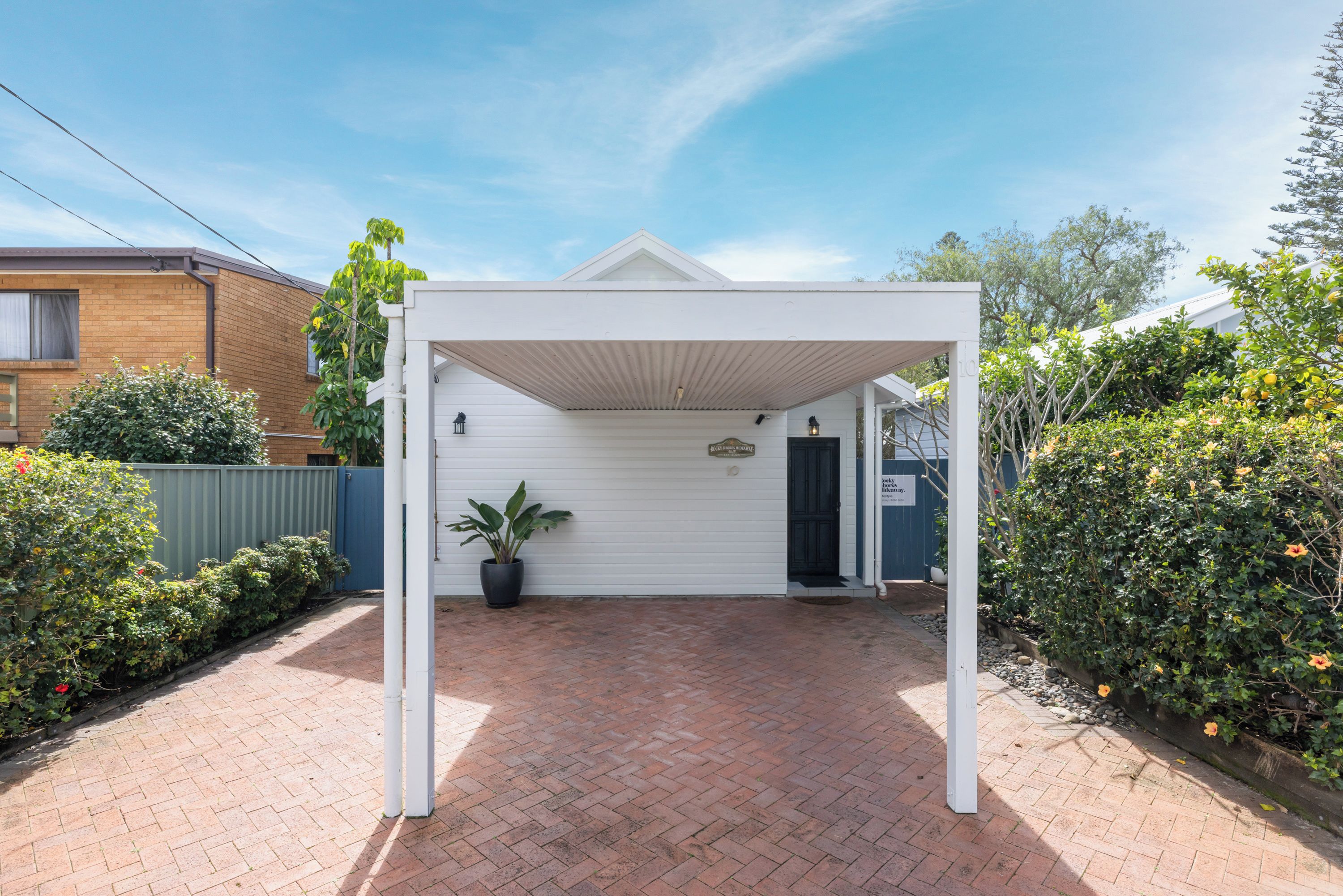 A covered patio area with a brick floor, surrounded by greenery and a white fence.