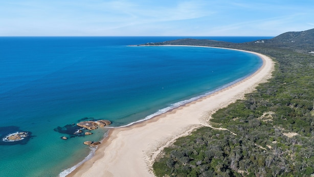 Luchtbeeld van een strand met helderblauw water, een zandstrand en weelderige begroeiing aan de oever.