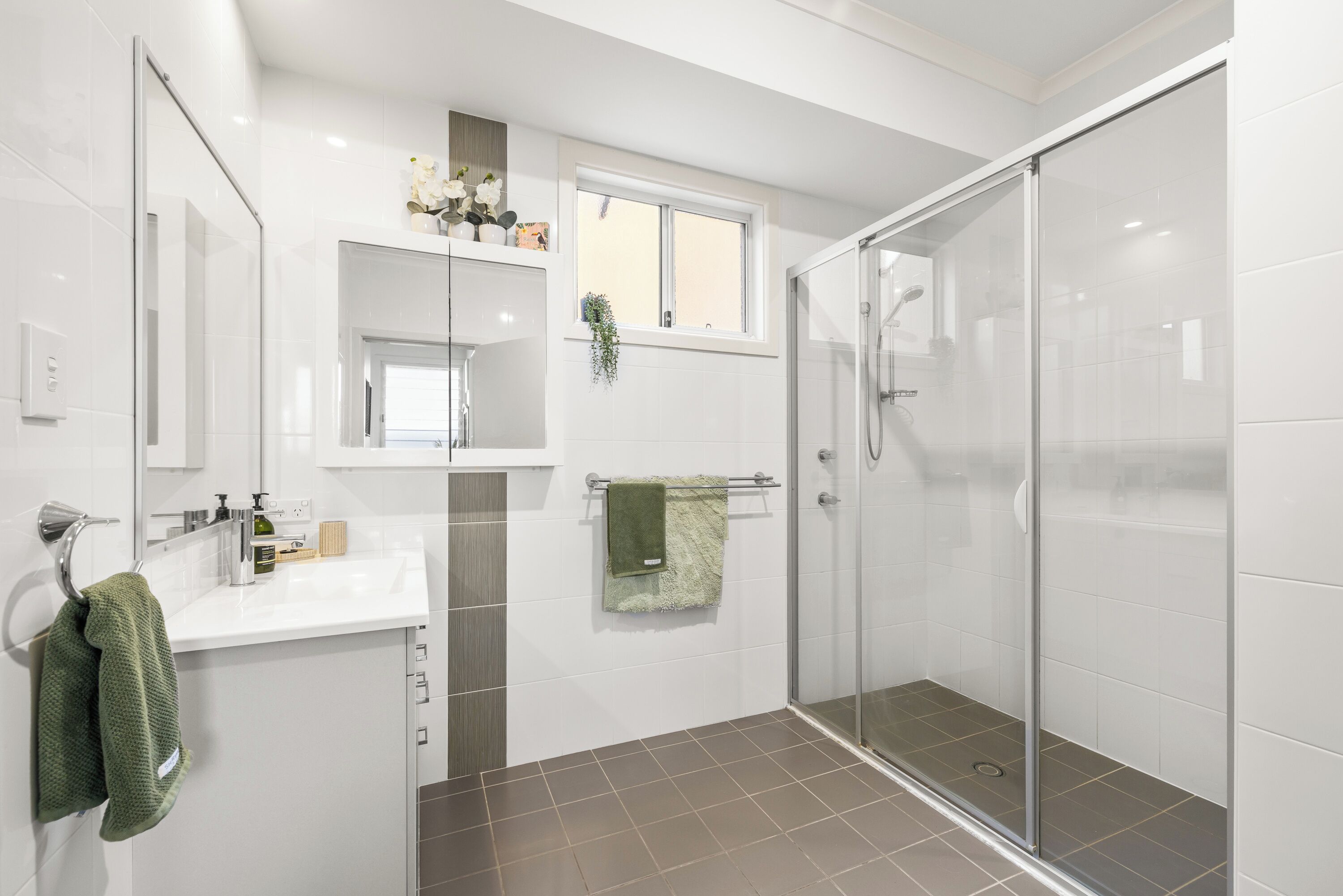 A modern bathroom with a glass shower enclosure, a white sink with a mirror above, and a green towel hanging on the wall.