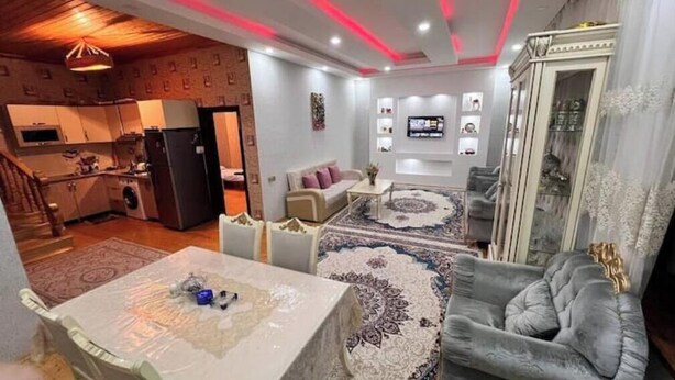A modern living room with a grey sofa, a coffee table, and a patterned rug. There is a kitchen in the background with a refrigerator and a washing machine. - Gabala Dreams, charming 2 bedroom villa, (Vandam)