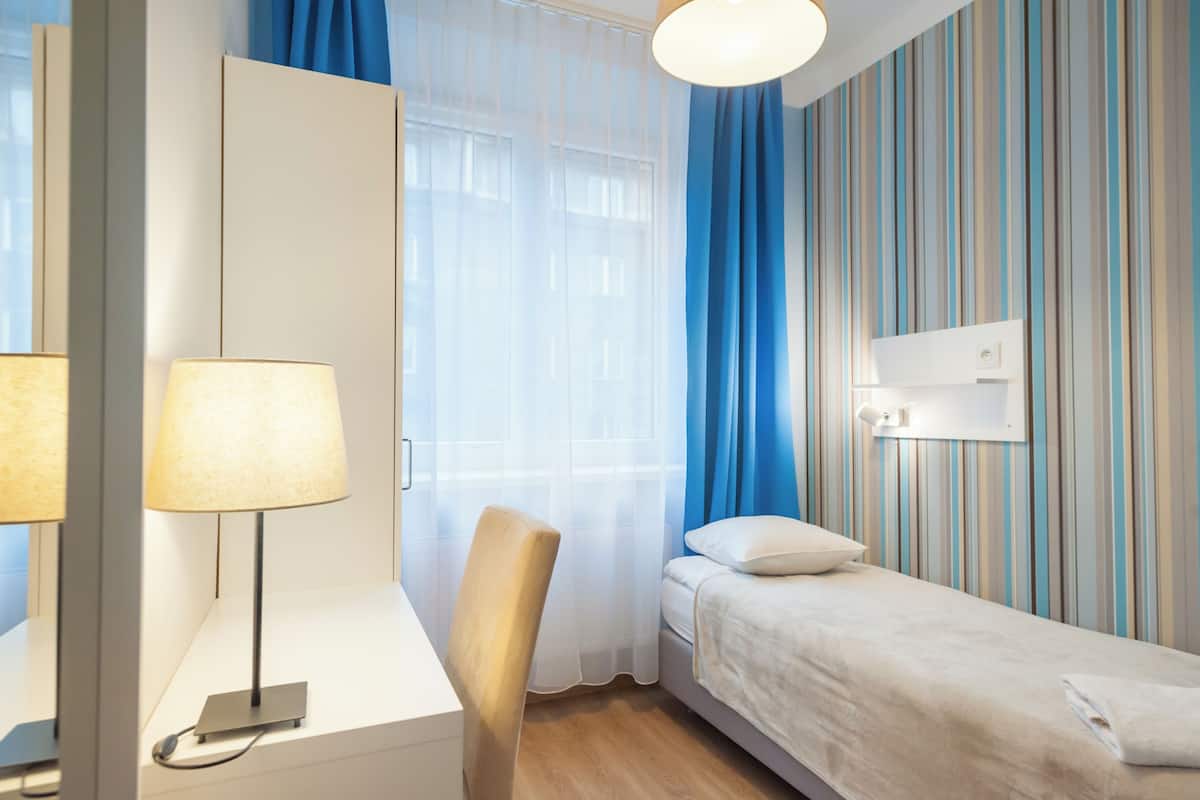 Ein Hotelzimmer mit einem Bett, einem Schreibtisch mit Lampe, einem Stuhl, einem Fenster mit durchsichtigen Vorhängen und einer gestreiften Wand.