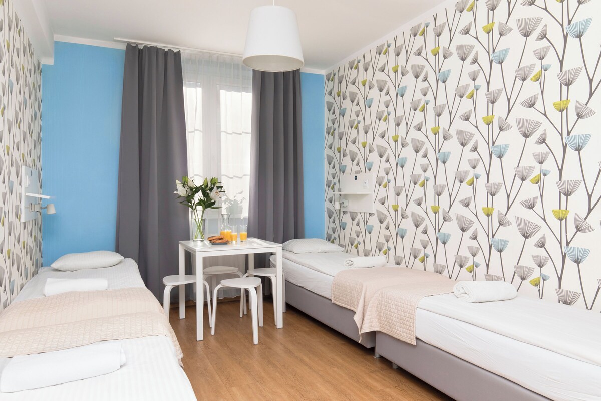 A modern hotel room with a bed, a small desk with a chair, a table with a vase of flowers, and a window with curtains.