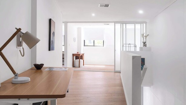 Un intérieur moderne avec un bureau en bois, une lampe, un tableau encadré, une petite table avec un coussin et une vue à travers une porte vitrée.