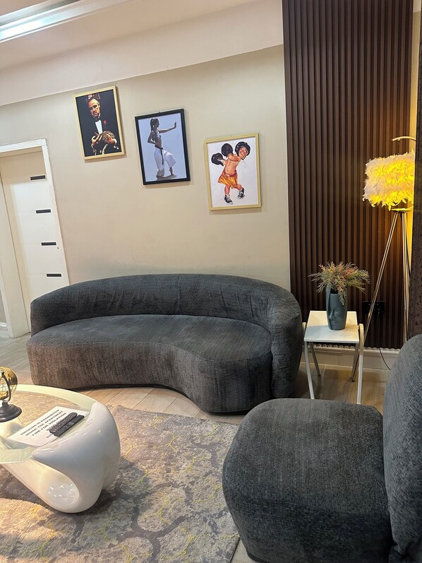 A modern living room with a curved grey sofa, a small white side table with a vase, and a floor lamp with a yellow shade. - majestic villa residence (Abuja)
