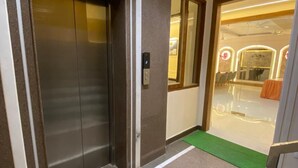 Een lift met een digitaal display, een groene mat bij de ingang en uitzicht op een ruimte met stoelen en een tafel.