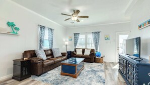 A living room with a brown leather sofa, a glass coffee table, a ceiling fan, and a TV on a black cabinet. - Surfside Beach Getaway Steps from the Ocean (Surfside Beach)