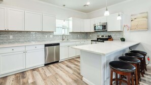 A modern kitchen with white cabinets, a stainless steel dishwasher, a microwave, and a large island with bar stools. - Surfside Beach Getaway Steps from the Ocean (Surfside Beach)