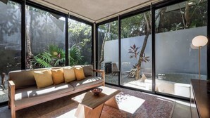 A modern interior with a sofa, a coffee table, and large windows offering a view of a garden and outdoor seating area.
