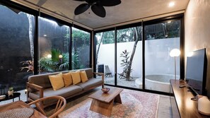 Un salon moderne avec un canapé, une table basse et des fauteuils, de grandes fenêtres donnant sur un jardin et un espace extérieur aménagé pour s’asseoir.