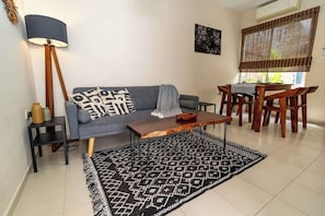 Uma sala de estar com um sofá cinza, uma mesa de centro de madeira, um tapete estampado, um abajur e uma área de jantar com cadeiras e mesa de madeira.