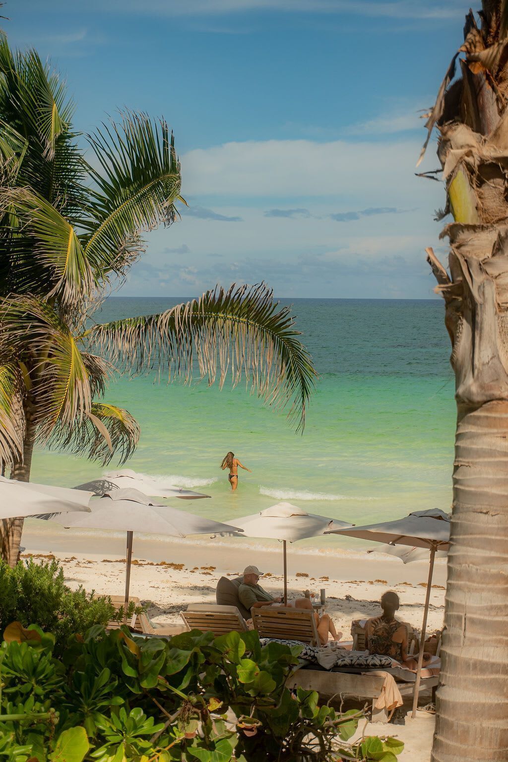 Een strand met helder turquoise water, palmbomen en strandstoelen.