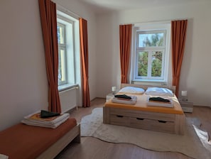 Ein Schlafzimmer mit zwei Betten, einem Heizkörper, einem Fenster mit Vorhängen, einem Nachttisch mit Lampe und einem Teppich.