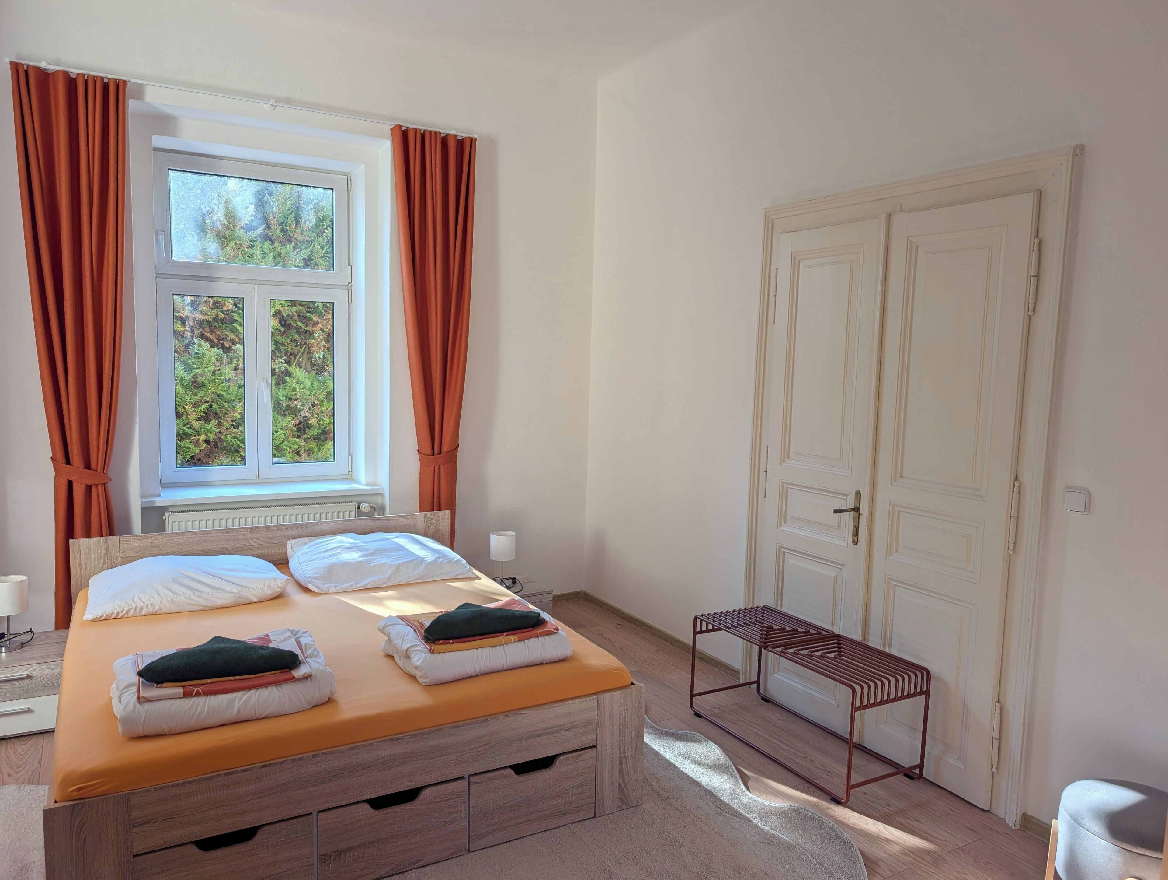 Ein Schlafzimmer mit Bett, Fenster mit Vorhängen, Heizkörper, Tür und einer Bank.
