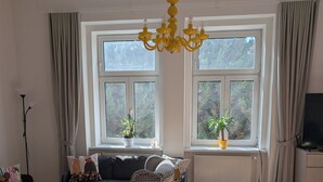 Een woonkamer met een bank, fauteuil en een gele tafel. Twee ramen met gordijnen, een radiator en een tv op een standaard.