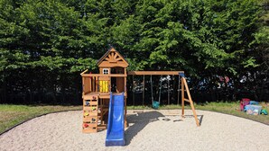 Een houten speelconstructie met een glijbaan en schommels, omringd door een grindgebied en bomen.