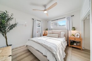 Une chambre à coucher avec un lit, un ventilateur de plafond, une fenêtre avec des rideaux, une table de chevet avec une lampe et une plante en pot.