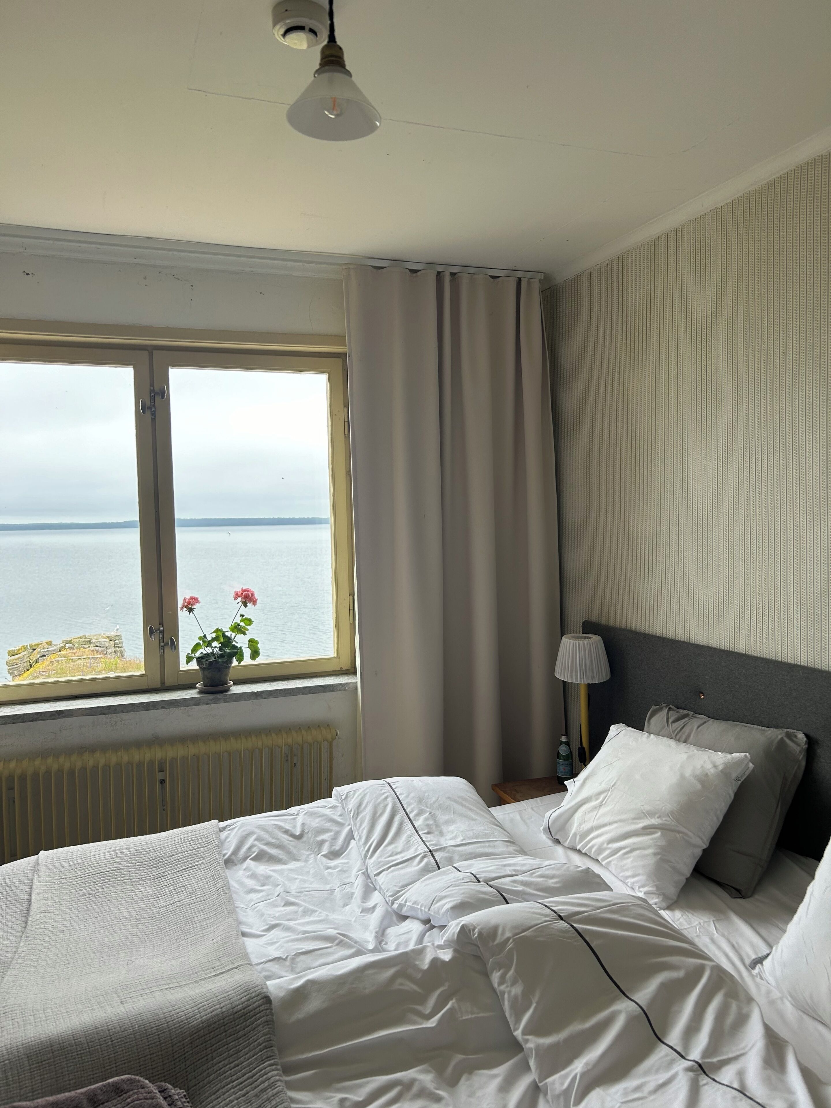 A bedroom with a bed, a window with curtains, a lamp, and a view of the sea.