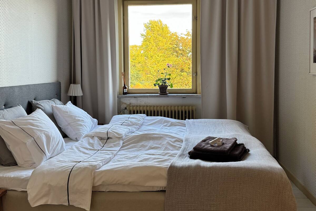 Una cama bien hecha con sábanas blancas, una lámpara de noche y una ventana con cortinas.