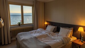 A hotel room with a large bed, two bedside lamps, a view of the sea through a window, and a radiator under the window. - Bungenäs Badhotell (Fårösund)