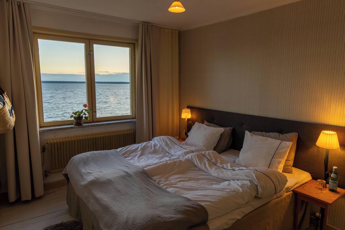 Un cuarto de hotel con una cama grande, dos lámparas de noche, vista al mar a través de una ventana y un radiador debajo de la ventana.