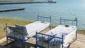 Two blue metal beds with striped bedding on a wooden deck by a calm body of water, with a distant boat and pier visible. - Bungenäs Badhotell (Fårösund)