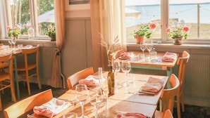 A dining area with tables set for two, large windows with curtains, and potted plants on the windowsill. - Bungenäs Badhotell (Fårösund)