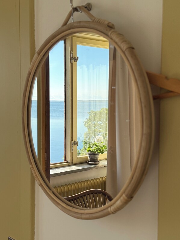 Habitación con un espejo ovalado grande que refleja la vista del mar a través de la ventana y una planta en maceta en el alféizar.