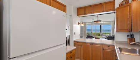 Una cocina con armarios de madera, un refrigerador blanco, una ventana con vista al océano y una lámpara colgante.