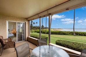 Een balkon met een glazen deur die uitkomt in een kamer, een ronde glazen tafel, rieten stoelen en uitzicht op een strand en palmbomen.